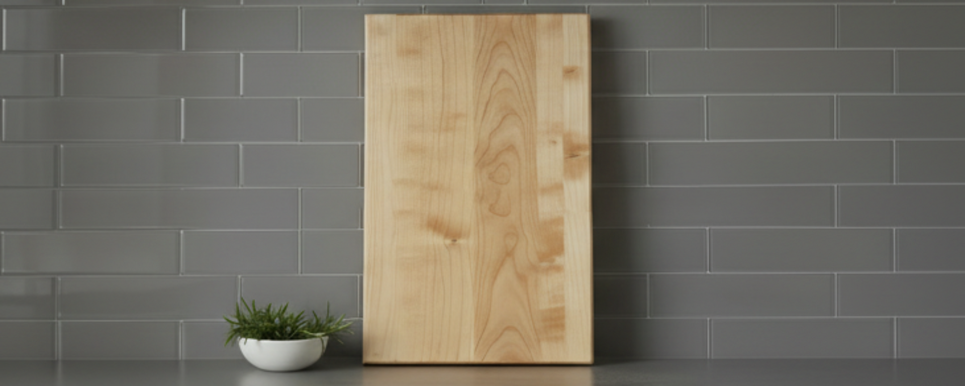 dark grey counter showing a maple cutting board with a background of grey subway tile backsplash, very minimalist kitchen.    need to have more width, reomve the black cylinder with utiensils
